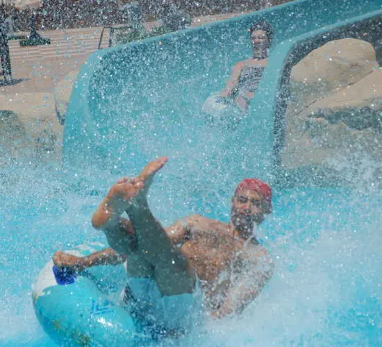 Colorful raft slide with riders splashing down the bright blue tunnel into the pool at Sharm El Sheikh’s aqua park, surrounded by palm trees and sunny skies.