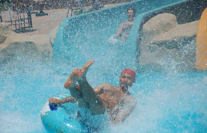 Colorful raft slide with riders splashing down the bright blue tunnel into the pool at Sharm El Sheikh’s aqua park, surrounded by palm trees and sunny skies.