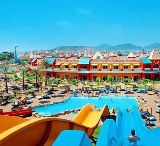 Massive green and orange open-top water slide with riders speeding down into the splash pool at Sharm El Sheikh's aqua park, surrounded by palm trees and sunny blue skies.