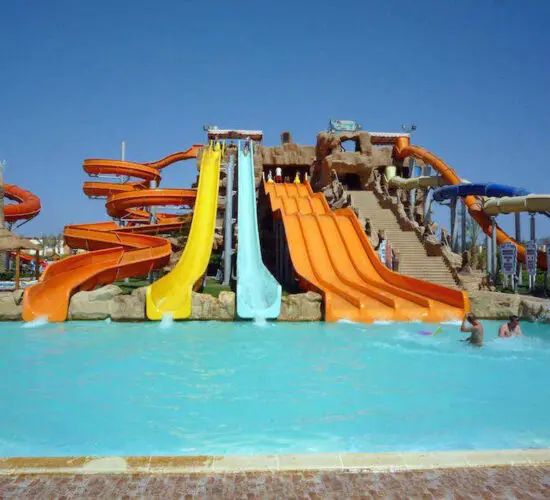 Thrilling multi-color water slides twisting down into the turquoise pool at Sharm El Sheikh's aqua park, with rocky themed structures and palm trees under sunny skies.
