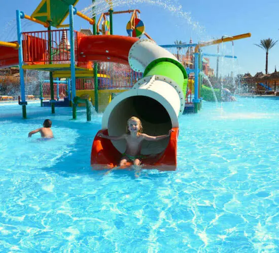 Joyful child splashing out of a colorful red water slide into the sparkling pool at Sharm El Sheikh’s family water park, with vibrant green and yellow play structures under sunny skies