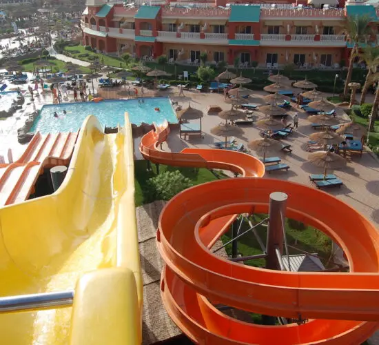 Aerial view of twisting yellow and orange water slides cascading into pools at Sharm El Sheikh's vibrant water park, surrounded by sun loungers, palm trees, and resort buildings under sunny skies.